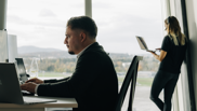 Frau steht telefonierend am Fenster und männlicher Microsoft 365 Architect sitzt im Vordergrund an PC