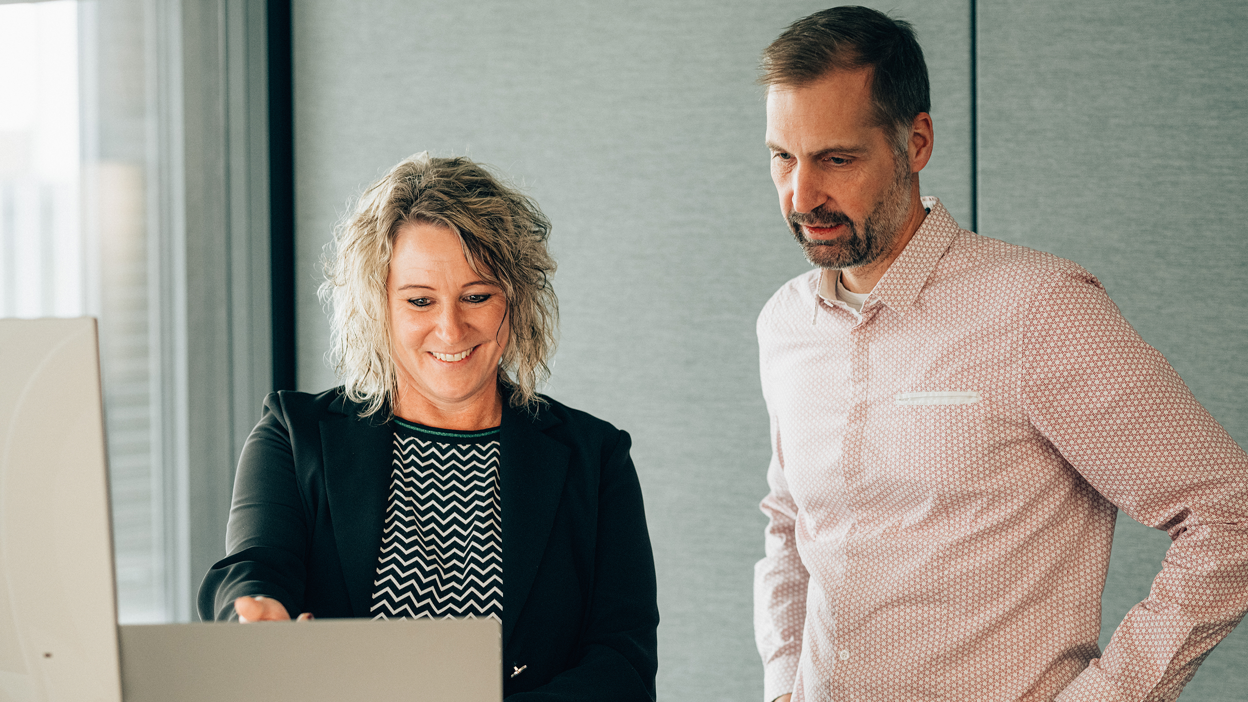 Ein männlicher Senior IT Strategy Consultant steht vor Laptop und hört Frau zu 