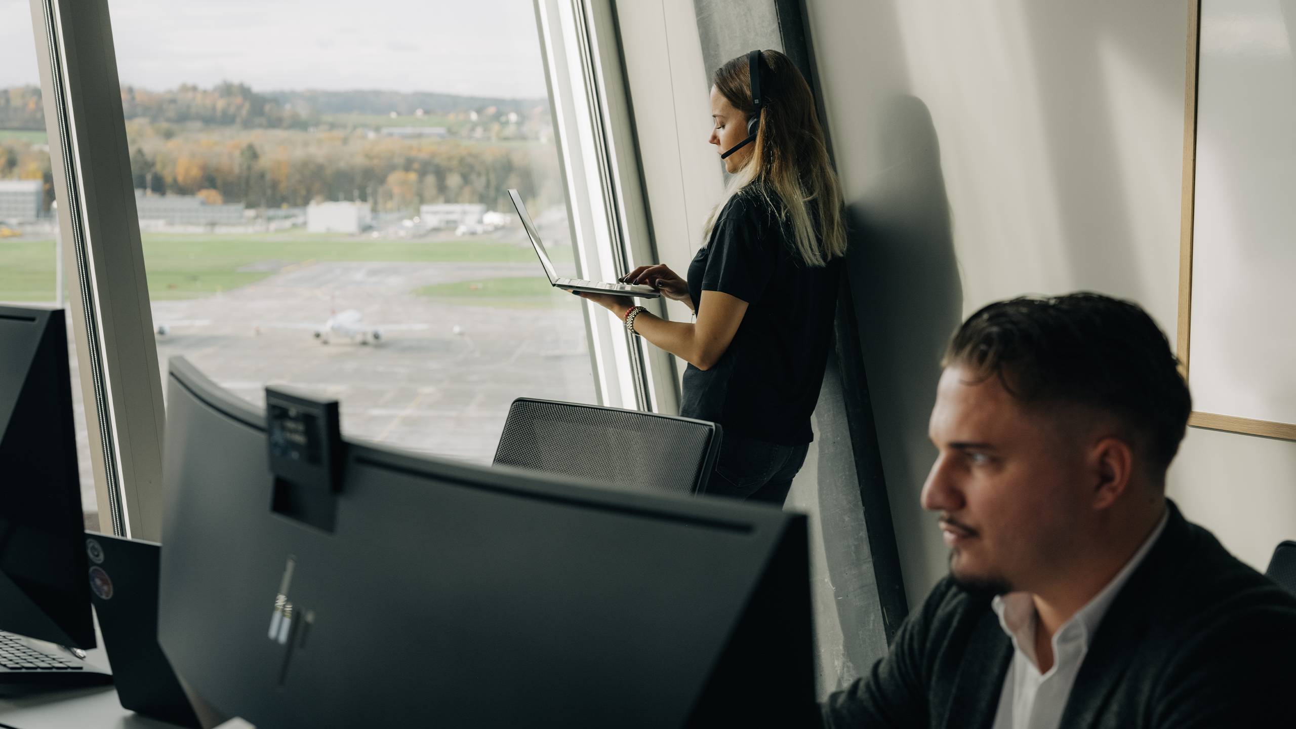 Man sitzt am PC im Hintergrund junge Dame am Laptop mit Headset vor Fensterfront