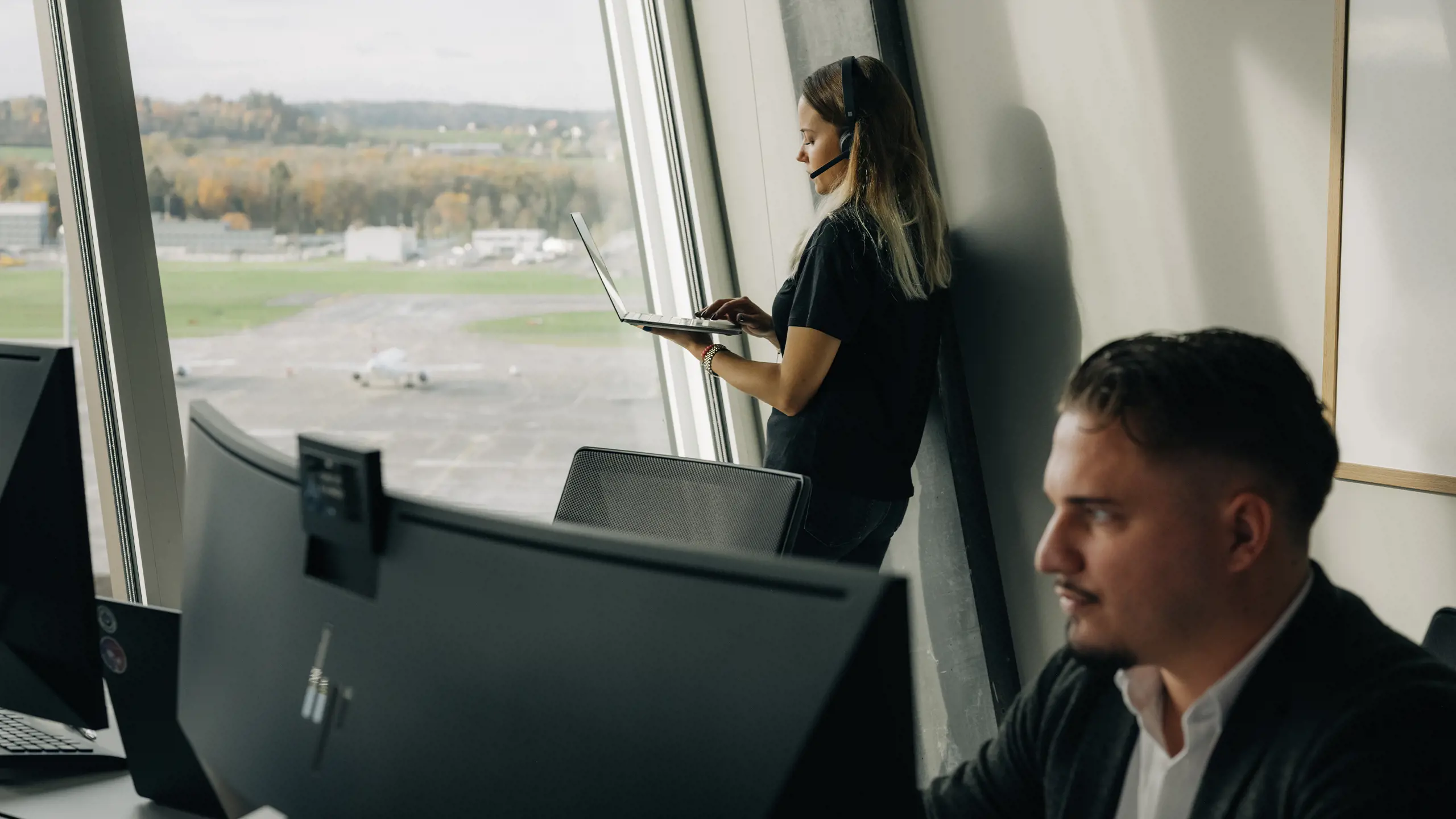 Man sitzt am PC im Hintergrund junge Dame am Laptop mit Headset vor Fensterfront