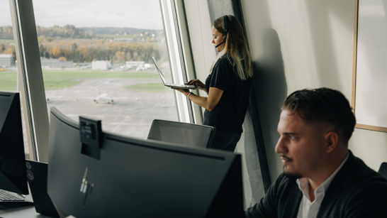 Application Security Man sitzt am PC im Hintergrund junge Dame am Laptop mit Headset vor Fensterfront