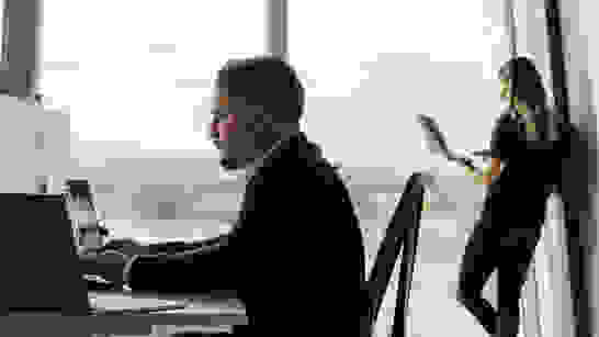Cloud Security Links Mann am PC und Frau hinten am Fenster angelehnt mit Laptop und Headset