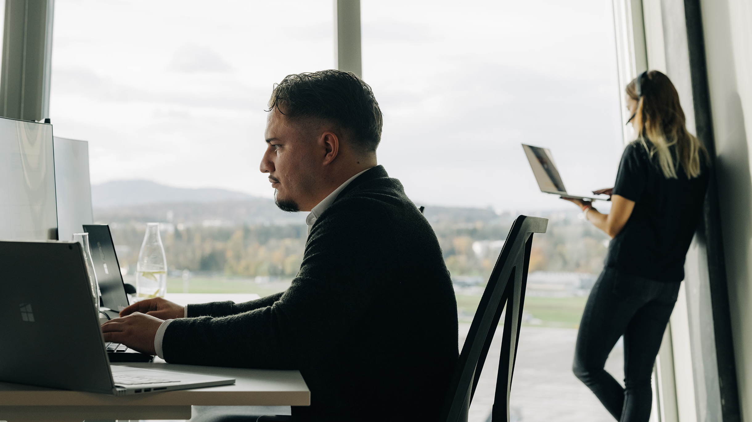 Links Mann am PC und Frau hinten am Fenster angelehnt mit Laptop und Headset