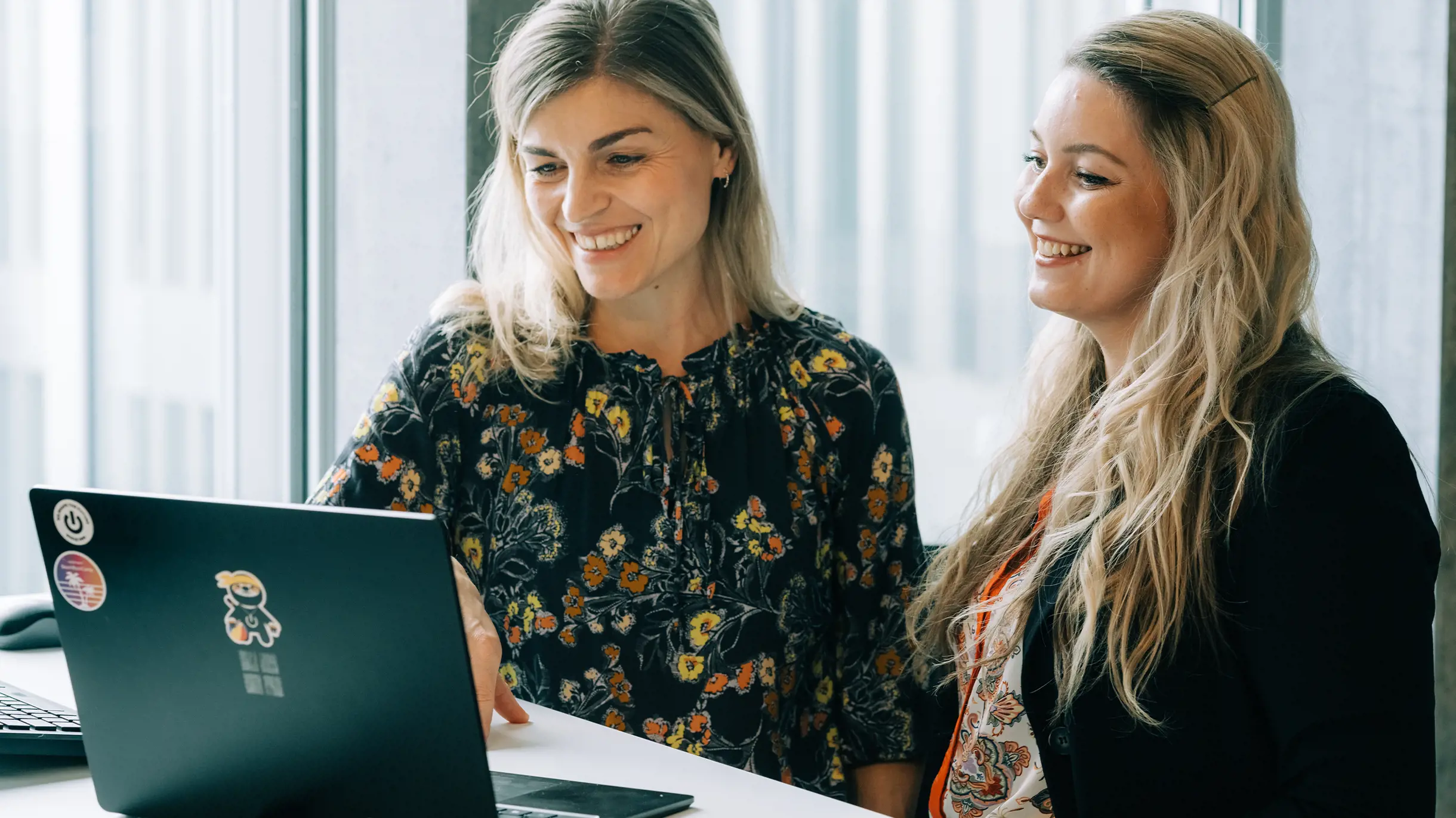 Zwei blonde Frauen vor einem schwarzen Surface Laptop lachend am reinschauen
