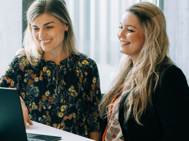 Zwei blonde Frauen vor einem schwarzen Surface Laptop lachend am reinschauen