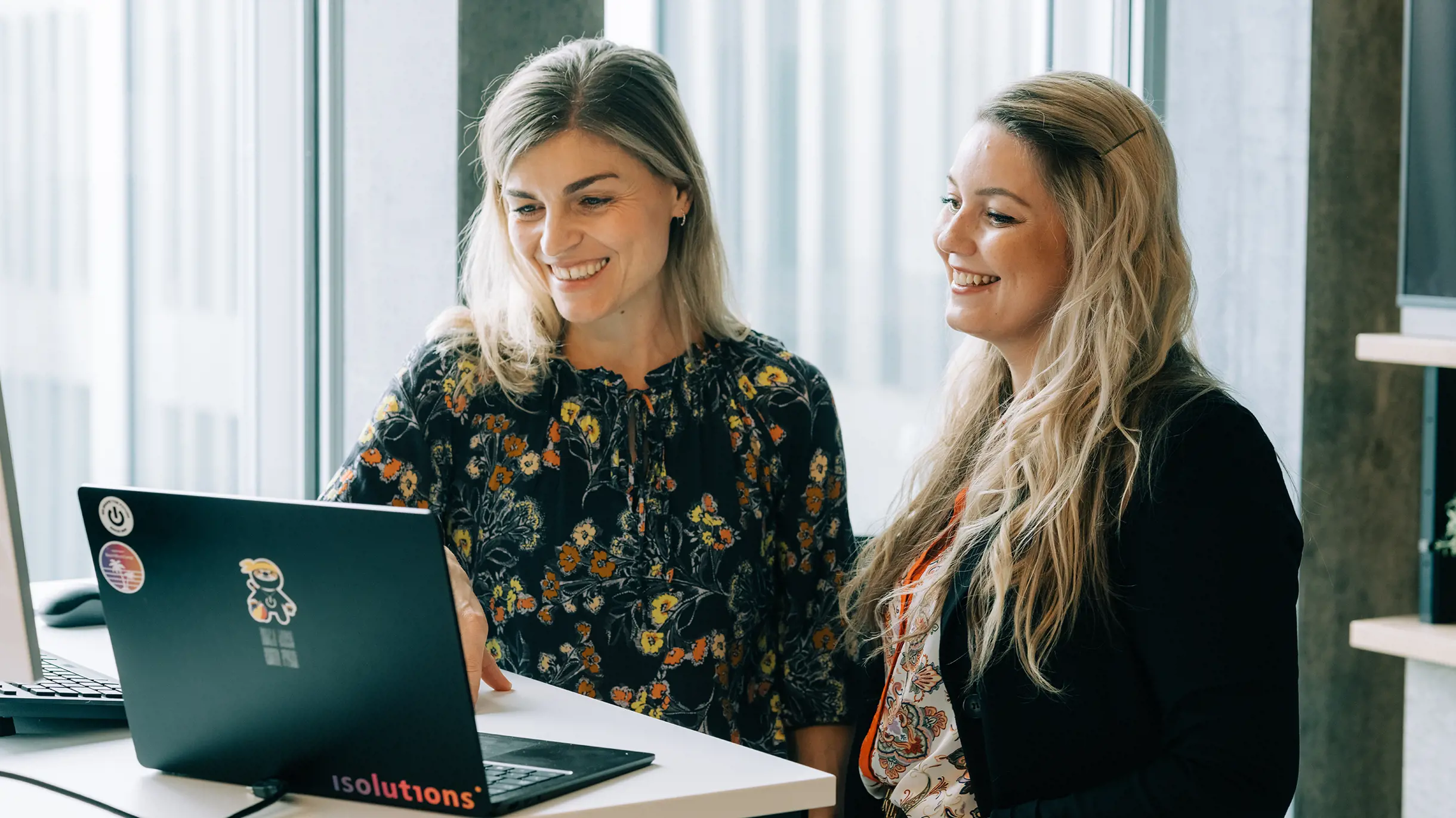 Zwei weibliche Backoffice Manager unterhalten sich vor Laptop und lachen dabei
