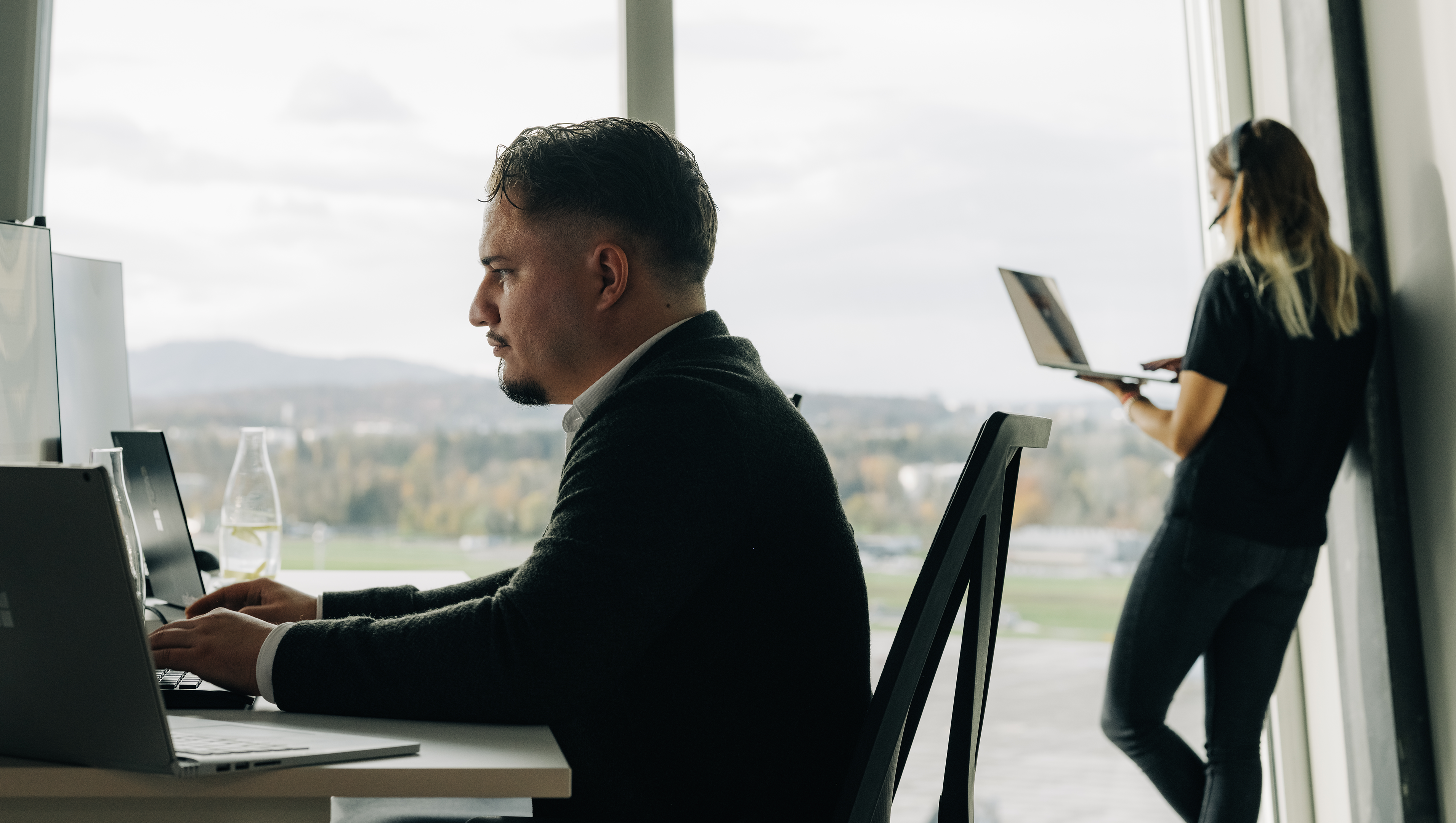 Frau steht telefonierend am Fenster und männlicher Microsoft 365 Architect sitzt im Vordergrund an PC