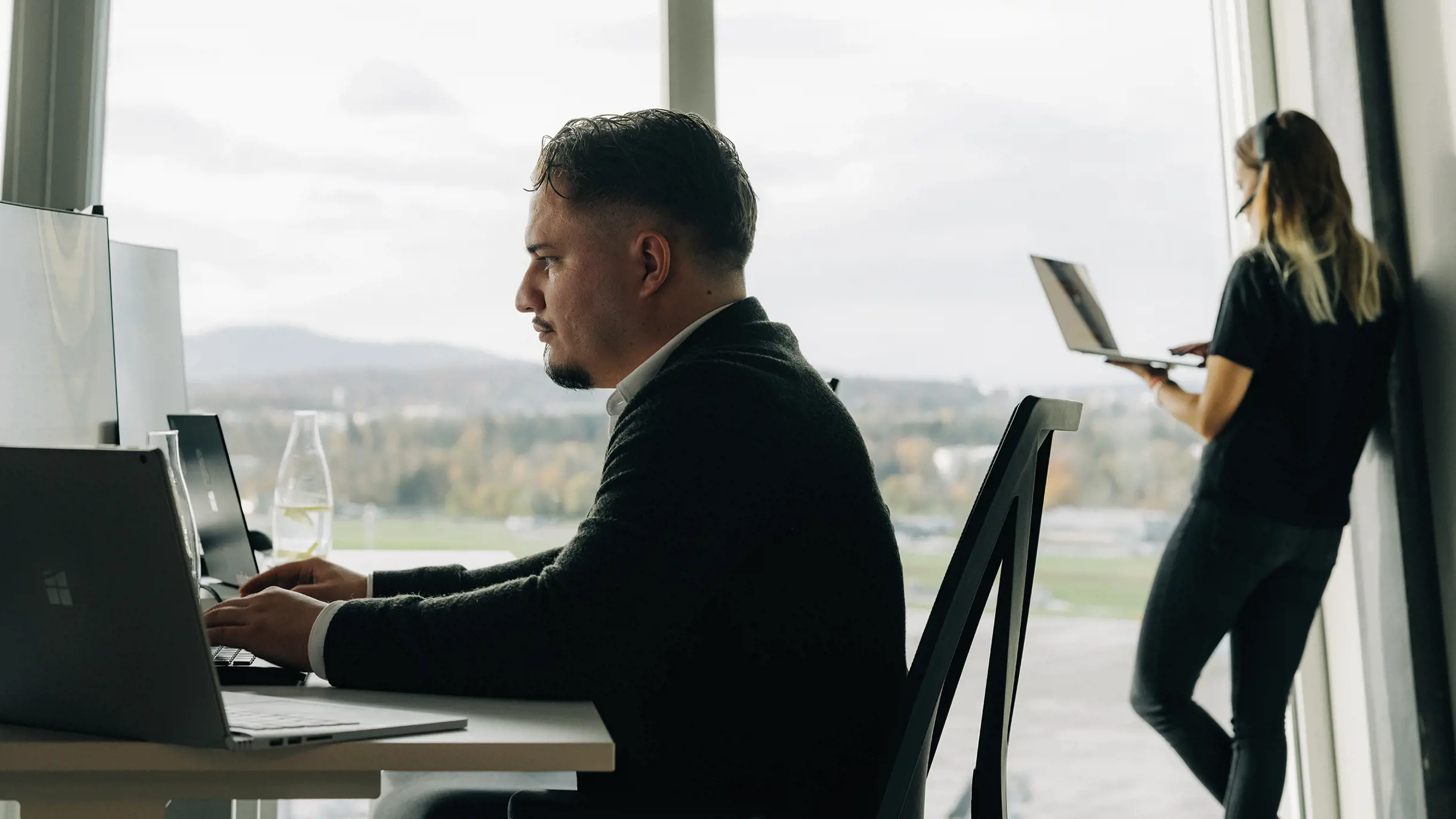 Frau steht telefonierend am Fenster und männlicher Project Manager sitzt im Vordergrund an PC