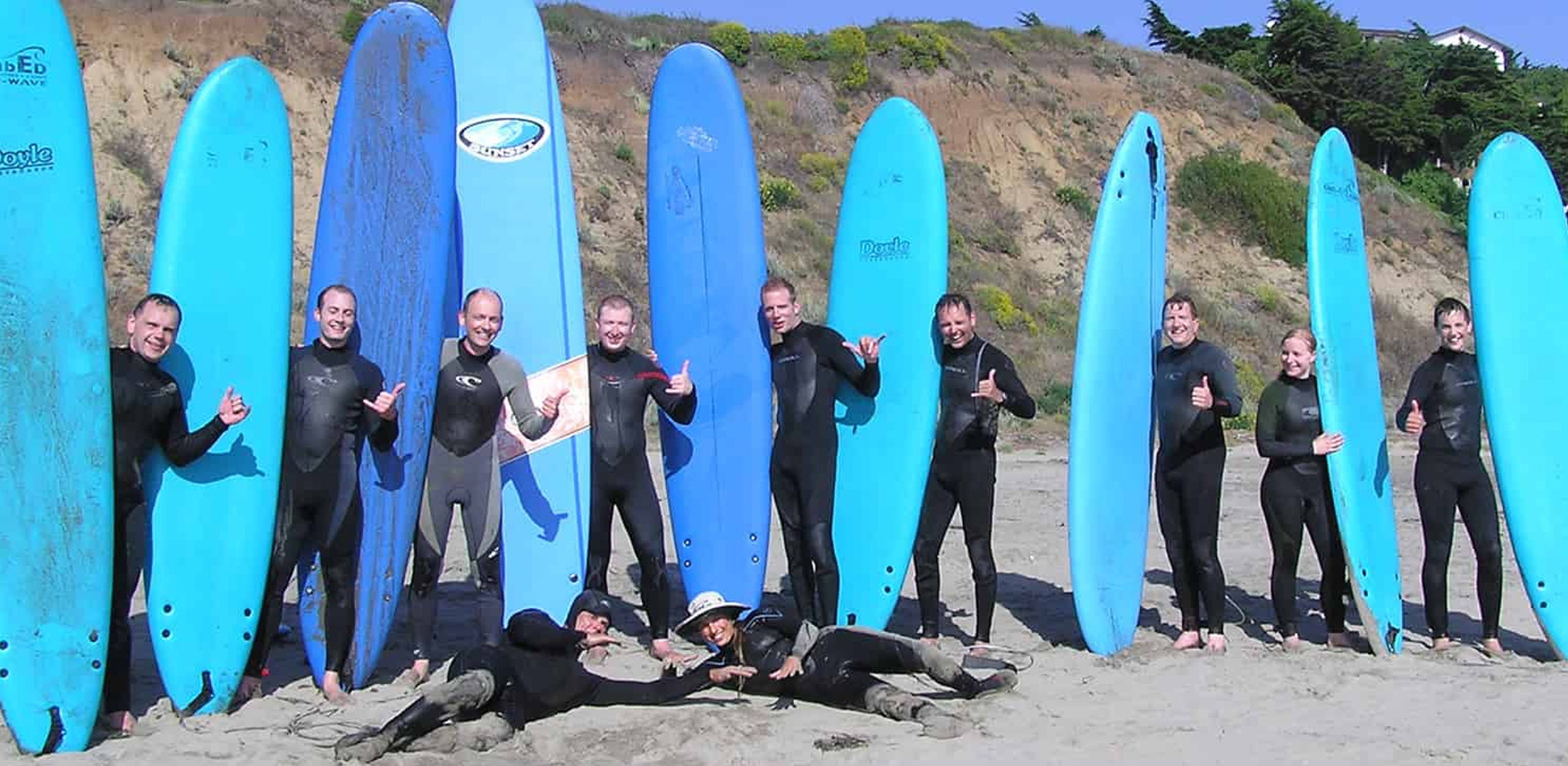 Sufer stehen nebeneinander und halten alle ein blaues Surfboard